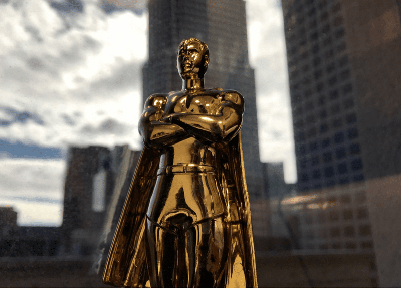 Golden statue of figure with cape standing against backdrop of city